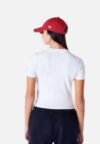 Witte T-shirt met korte mouwen en een getailleerd ontwerp, gecombineerd met zwarte broek. Rode pet met een gebogen rand, voorzien van een logo aan de zijkant.