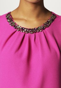 Fuchsialinne med veckad halsringning prydd med gröna och lila smyckeaccent, tillverkad av mjukt, texturerat tyg.