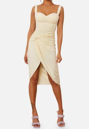 Femme portant une robe crème sans manches avec un nœud torsadé et une fente à l'avant, assortie à des talons lilas à brides.