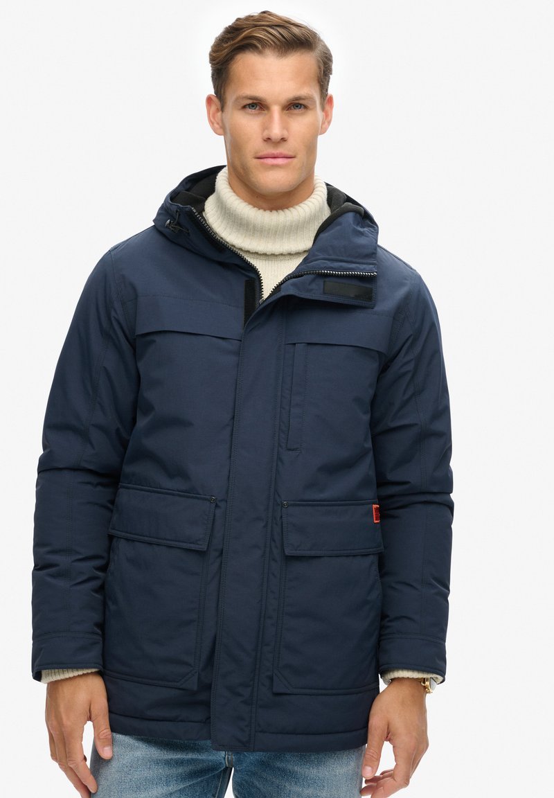 Marineblauwe geïsoleerde parka met een ritskap, twee voorzakken en een gebreide kraag. Voorzien van een stevige rits en klittenbandaccenten.