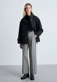 Veste en laine noire avec fermetures à pression, pantalons gris taille haute et bottines noires. Design minimaliste et silhouette structurée.