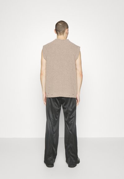 Άτομο με κοντά μαλλιά που φοράει μια μπεζ αμάνικη πλεκτή μπλούζα και φαρδιά μαύρα σατινέ παντελόνια, στέκεται μπροστά σε έναν απλό άσπρο τοίχο.