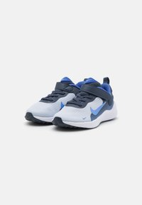 Baskets Nike avec une tige en mesh gris clair, des accents bleu marine, un swoosh bleu et une sangle Velcro pour un maintien sécurisé. Semelle intermédiaire blanche, semelle texturée.