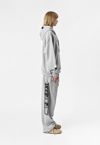 Sudadera con capucha de color gris claro y joggers a juego. Sudadera con bolsillo frontal, joggers que presentan gráficos impresos en negro en el costado.