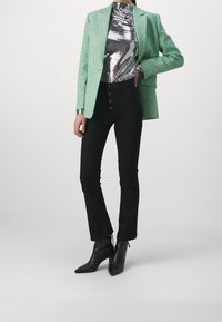 Zelený tvídový blejzr přes lesklý stříbrný top, spárovaný s černými kalhotami s vysokým pasem a širokými nohavicemi a černými špičatými kotníkovými botami.