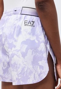 Κοντινό πλάνο ατόμου που φοράει ανοιχτό μωβ και λευκό σορτς με μοτίβο και λογότυπο EA7 Emporio Armani στο πλάι και λευκό πουκάμισο.