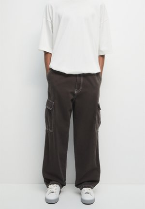 Personne portant un pantalon cargo noir ample avec des surpiqûres blanches contrastantes, une chemise blanche oversize à manches courtes et des baskets blanches, les mains dans les poches.