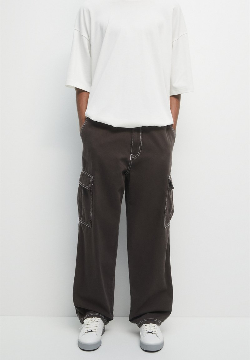 Personne portant un pantalon cargo noir ample avec des surpiqûres blanches contrastantes, une chemise blanche oversize à manches courtes et des baskets blanches, les mains dans les poches.