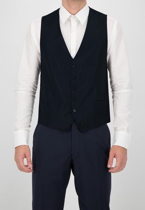 Homme portant une chemise blanche avec un gilet bleu marine boutonné et un pantalon de costume bleu marine assorti, debout devant un fond uni.