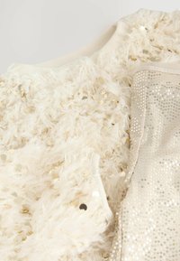Weißer flauschiger Pullover mit goldenen Pailletten auf der Vorderseite, kombiniert mit glänzenden beigen Hosen, die kleine runde Pailletten auf dem Stoff haben.