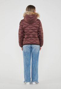 Chaqueta acolchada marrón con diseño de rombos, capucha con borde de piel, y acentos en negro, llevada sobre jeans de pierna ancha azul claros y zapatillas blancas.