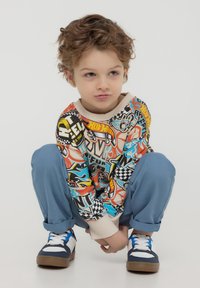 Felpa grafica colorata con stampe a tema corsa, polsini beige, abbinata a pantaloni blu e sneakers navy, seduto in posizione accovacciata.