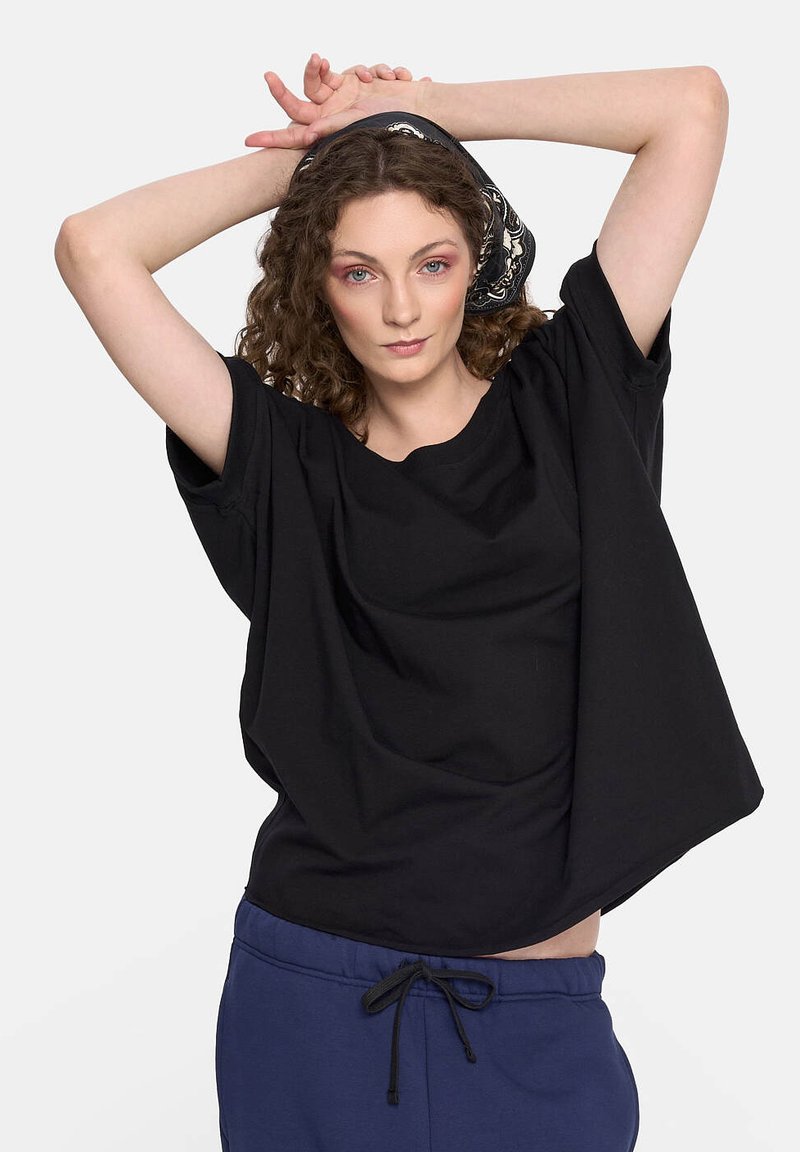 Luźna czarna koszulka bawełniana z szerokimi rękawami i okrągłym dekoltem. Modelka nosi wzorzystą czarno-białą chustę na głowie. Styl casualowy z luźnym krojem.