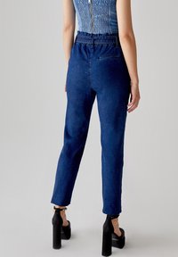 Jeans in denim di colore blu scuro, a vita alta con gamba affusolata. Presentano tasche posteriori e una texture liscia, abbinati a décolleté con tacco a piattaforma neri.