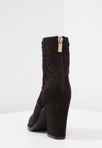 Bota de tobillo negra de tacón alto hecha de ante con detalles trenzados. Presenta un cierre lateral y un diseño de punta redonda.