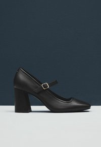 Černé kožené Mary Jane boty s blokovým podpatkem a malou zlatou sponou, umístěné na tmavě modrém a bílém pozadí.