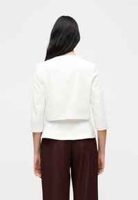 Femme aux longs cheveux foncés vue de dos, portant une veste courte blanche à manches trois-quarts et un pantalon brun foncé, sur un fond uni.