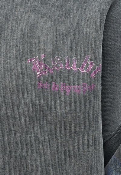 Sivé látkové pozadie s fialovým vyšívaným textom v gotickom písme s nápisom "Ksubi" a menším textom pod ním na odevu.