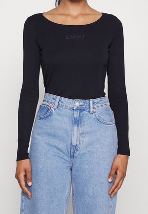Frau trägt schwarzen Langarm-Top mit "ESPRIT" Strass-Logo und hellblauen hochgeschnittenen Jeans, die Hände entspannt an den Seiten.