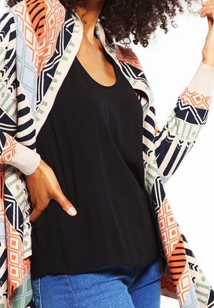 Nainen pukeutuneena mustaan yläosaan, sinisiin farkkuihin ja moniväriseen, geometrisesti kuvioituun neuletakkiin, yksi käsi taskussa ja näkyvissä kiharat hiukset.