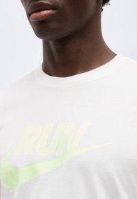 Κοντινό πλάνο ενός ατόμου που φοράει λευκό t-shirt με αχνό πράσινο κείμενο "RUN" και λογότυπο swoosh στο στέρνο.