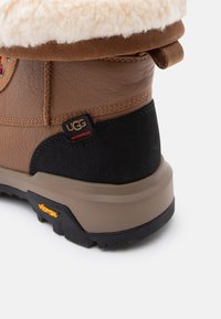 Bruine waterdichte laars met een beige suède boord, voering van schapenvacht, rubberen zool en zwart textuurpaneel. Voorzien van UGG-logo.