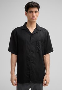 Schwarzes Kurzarmshirt aus leichtem Stoff mit spitzem Kragen, Fronttasche und Knopfleiste. Glatte Textur, lockere Passform.