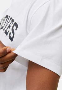 Hvid bomulds t-shirt med korte ærmer, med en dristig sort grafisk tekst på forsiden. Nærbillede af en hånd, der justerer ærmet.