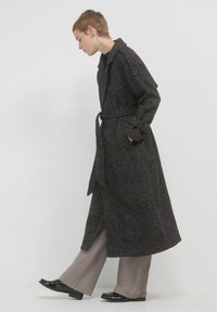 Lungo cappotto strutturato nero e grigio con cintura, tasche laterali e colletto. Abbinato a pantaloni grigi chiari a gamba larga e scarpe nere lucide.