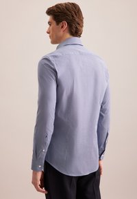 Man die zijwaarts staat, naar links gericht, in een lichtblauwe lange mouw blouse en zwarte broek tegen een effen achtergrond.