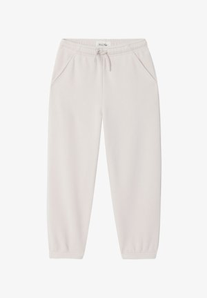 Junge steht und trägt ein rotes „American Vintage“-T-Shirt, weiße Hosen und beige Sandalen vor einem schlichten hellen Hintergrund.