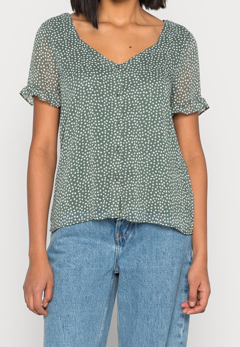 Kvinna i grön kortärmad blus med vita prickar och volangmuddar, tillsammans med ljusblå högmidjade jeans.