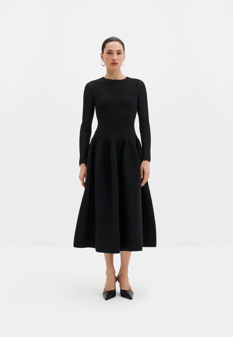 Femme debout portant une robe noire à manches longues avec un corsage plissé ajusté et une jupe évasée, associée à des talons noirs pointus et des boucles d'oreilles créoles dorées.