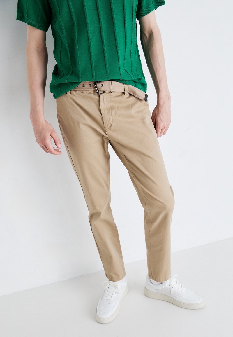 Beige bavlněné chinos s rovný fit, s jednoduchým designem předních kapes a hnědým opaskem. Spárováno s bílými teniskami.