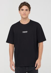 T-shirt nera in cotone con maniche corte, scollatura rotonda e testo bianco "CHICAGO RAW MUSIC SCENE" stampato sul petto.