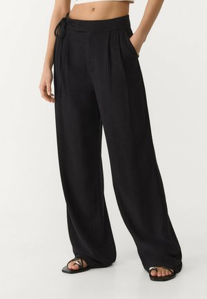 Vrouw draagt losse zwarte wijde broek met zijzakken en zwarte sandalen met open tenen, staand tegen een lichte achtergrond.