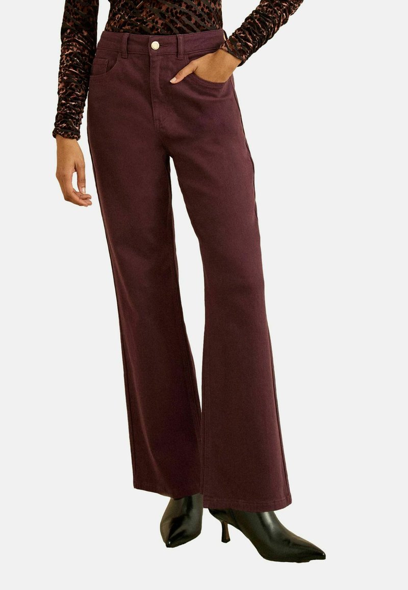 Burgundi värvi laienevad püksid siledast kangast, millel on esipockets ja nupuga kinnitamine. Sobitatud mustade teravate kingadega.