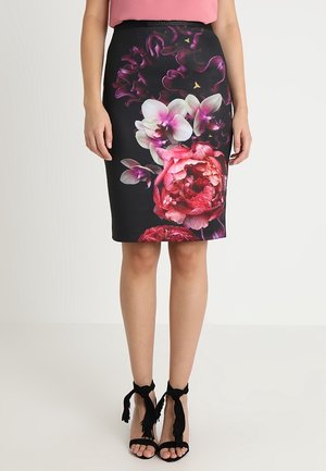 Falda lápiz negra hasta la rodilla con gran estampado floral en rosa y blanco, combinada con sandalias negras de tacón alto con lazos en el tobillo.