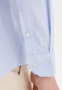 Jasnoniebieska lniana koszula z długimi rękawami, z zapięciem na guzik przy mankiecie i delikatną teksturą materiału. Białe guziki są widoczne na mankiecie.