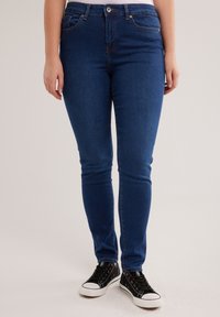 Partea inferioară a corpului unei persoane care poartă blugi skinny albastru închis și adidași negri cu talpă joasă, pe un fundal neutru.