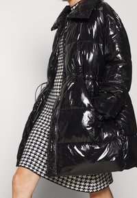 Manteau d'allaitement noir en duvet avec une finition brillante, doté d'un col haut et d'une taille ajustable avec cordon de serrage. Doublé d'un motif pied-de-poule.