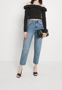 Svart off-shoulder topp med volanger, ihop med ljusblå högmidjade jeans och svarta klackskor. Håller en svart kuvertväska med guldhandtag.