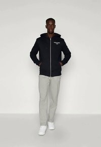 Črna zip-up kapuca z logotipom spredaj, v kombinaciji z svetlo sivo obleko in belimi teniskami. Model stoji s rokami v žepih na enotni podlagi.