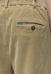Pantalon beige en velours côtelé avec poche arrière à fermeture bouton et petite étiquette bleue de la marque Barbour sur le bord de la poche.