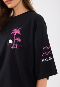Tricou negru cu un design în roz al unei palmieri și al unui soare pe față. Textul de pe mânecă spune "CĂUTAȚI SUB PALMA" în roz și alb.