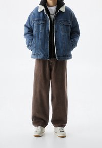 Persona con una chaqueta de mezclilla azul forrada de sherpa, sudadera con capucha negra, pantalones anchos marrones, zapatillas blancas y un collar de cadena plateada.