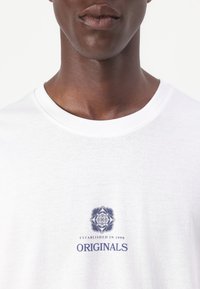 Camiseta blanca de algodón con escote redondo y un diseño gráfico azul que incluye el texto "Establecido en 1990" y "ORIGINALS."