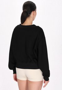 Svart sweatshirt med boxig passform och ribbade detaljer vid ärmar och nederkant. Modellen bär beiga shorts, sedd bakifrån.