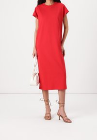 Rotes Midi-Kleid mit Kap sleeves und entspannter Passform, kombiniert mit beigen Riemchen-Heels und einer weißen Handtasche, vor einem schlichten Hintergrund.