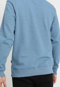 Světlě modrá mikina s kulatým výstřihem vyrobená z měkké bavlněné směsi. Má žebrované lemy na rukávech a spodním okraji a malou logo štítek na levé straně.